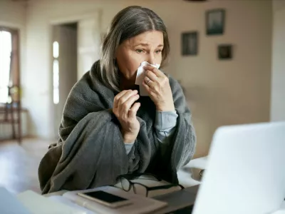 Sick elderly woman wrapped in plaid coughing, sneezing, having respiratory disease symptoms, sitting in front of open laptop, having online video consultation with her physician. Health and technology / Foto: Shurkin_son