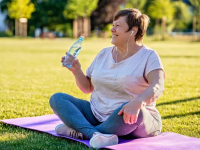 Relaxed senior plus size woman with earphones sitting on yoga mat on green grass outdoors resting after exercises drinking water at warm sunny summer day / Foto: Tetiana Soares