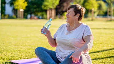 Relaxed senior plus size woman with earphones sitting on yoga mat on green grass outdoors resting after exercises drinking water at warm sunny summer day / Foto: Tetiana Soares