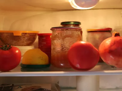 Food items, including bright tomatoes, a lemon, and a pomegranate, sit on a glass shelf inside a refrigerator next to homemade preserves in jars. This close-up shot illustrates food storage and supplies inside the kitchen appliance. / Foto: Oleh Strus Getty Images
