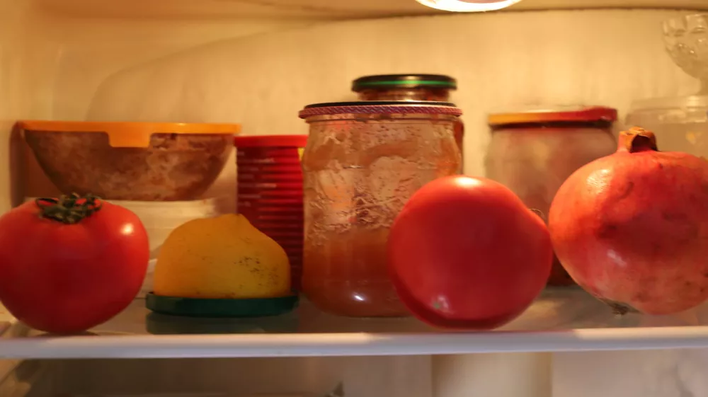 Food items, including bright tomatoes, a lemon, and a pomegranate, sit on a glass shelf inside a refrigerator next to homemade preserves in jars. This close-up shot illustrates food storage and supplies inside the kitchen appliance. / Foto: Oleh Strus Getty Images