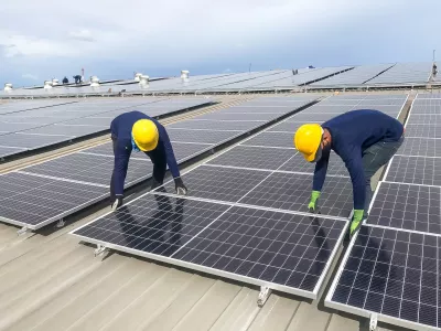 Male team workers in protective helmet installing solar photovoltaic panel system using screwdriver. Electrician mounting blue solar module on roof of modern house.