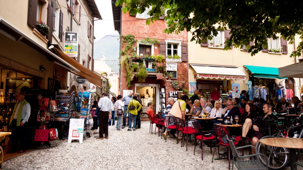 "Malcesine, Italy - April 29, 2012: Lot of people sitting in the cafe drinking and talking. Few people standing and shoping at the town sqare"