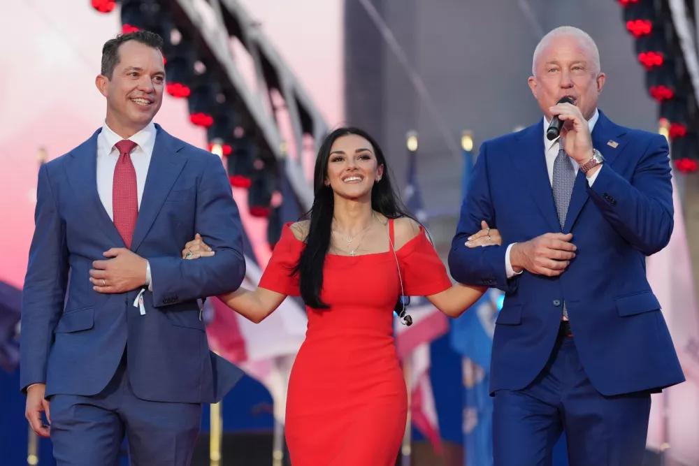 U.S. Ambassador to Belgium Bill White, right, and his husband Bryan Eure, left, walk U.S. country music singer Alexis Wilkins onto the stage during the America 250 event in Brussels, Sunday, June 28, 2026. (AP Photo/Virginia Mayo)