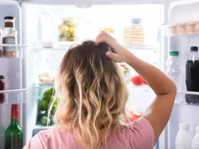 Rear View Of A Confused Woman Looking In Open Refrigerator At Home