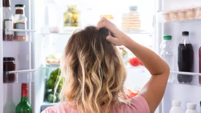 Rear View Of A Confused Woman Looking In Open Refrigerator At Home