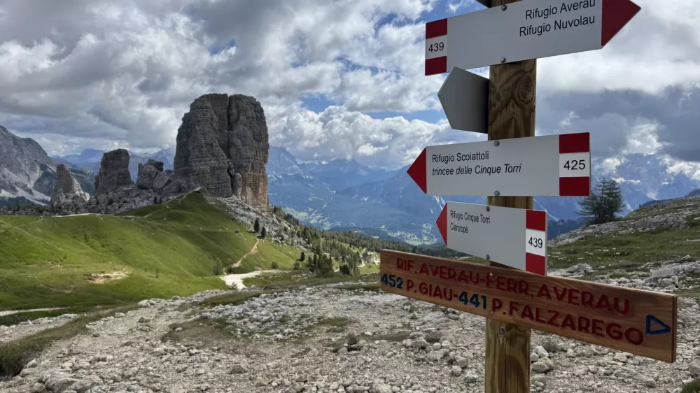 The photo captures a scenic mountain landscape with a rugged trail leading into the distance. Dominating the background are the striking rock formations of the Cinque Torri, a well-known landmark in the Dolomites of Italy. The sky is partly cloudy, casting dynamic shadows over the rocky and grassy terrain. In the foreground, there's a wooden signpost with multiple directional signs indicating hiking routes to destinations such as Rifugio Averau, Rifugio Nuvolau, Rifugio Scoiattoli, and Cinque Torri. The signs are marked with trail numbers 439 and 425, and the wooden plaque below provides additional trail information. The overall scene conveys a sense of adventure and natural beauty, ideal for hiking enthusiasts.