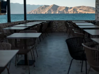 An empty outdoor dining area with tables and chairs is set up on a terrace overlooking the blue sea and distant rocky mountains