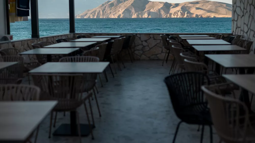 An empty outdoor dining area with tables and chairs is set up on a terrace overlooking the blue sea and distant rocky mountains