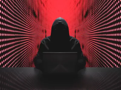 Anonymous hacker in front of his computer with red light wall backgroundAnonymous hacker in a black hoody with laptop in front of a code background with binary streams cyber security concept