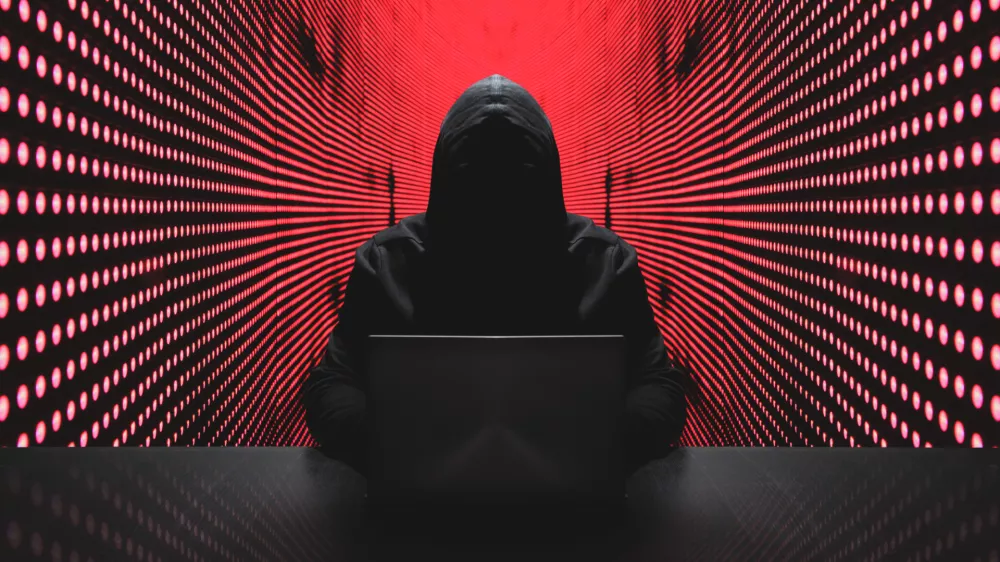 Anonymous hacker in front of his computer with red light wall backgroundAnonymous hacker in a black hoody with laptop in front of a code background with binary streams cyber security concept