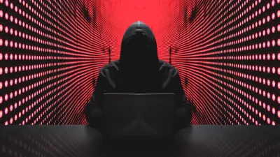 Anonymous hacker in front of his computer with red light wall backgroundAnonymous hacker in a black hoody with laptop in front of a code background with binary streams cyber security concept