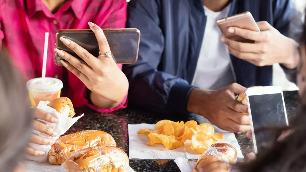 Group of unrecognizable people busy on mobile while having snacks together at restaurant - Concept of millennials having junk food, phone addiction, social media sharing and connection / Foto: Lakshmiprasad S Getty Images