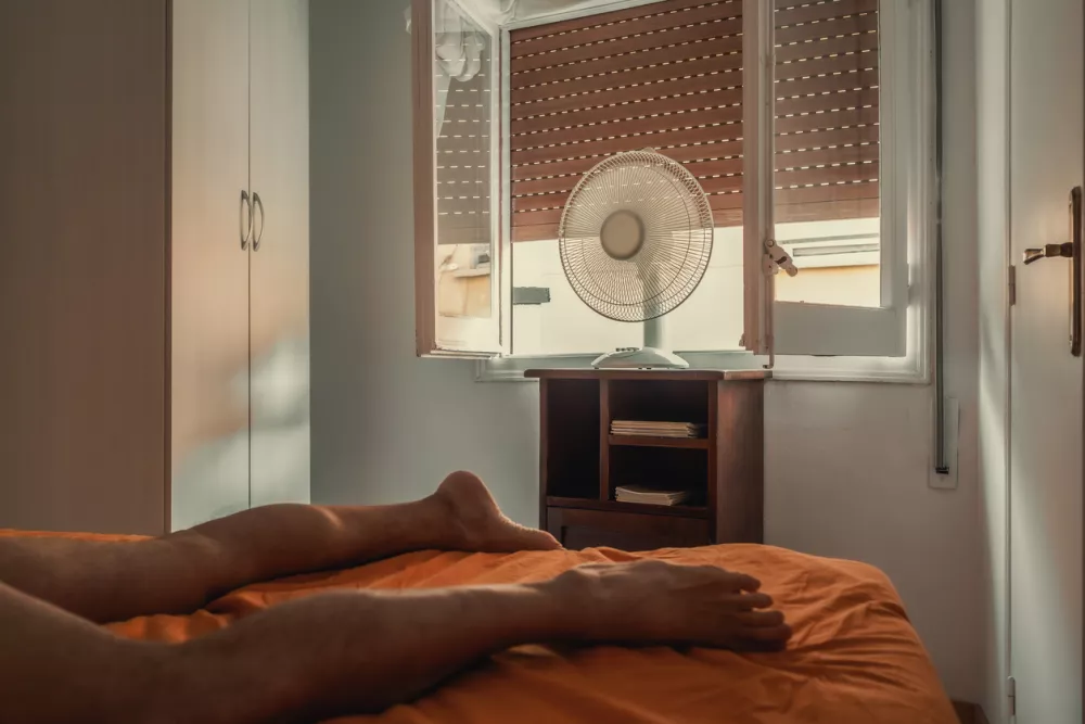 Man lying on a bed in a bedroom, struggling to cope with the sweltering summer heat wave. An electric fan positioned by the open window offers some relief from the rising temperature. / Foto: Riccardo Cirillo Getty Images