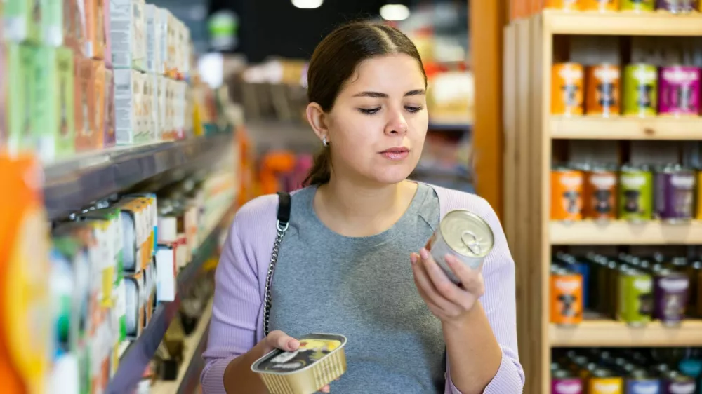 Positive young woman choosing canned natural nutritious dog food for her pet in store / Foto: Jackf