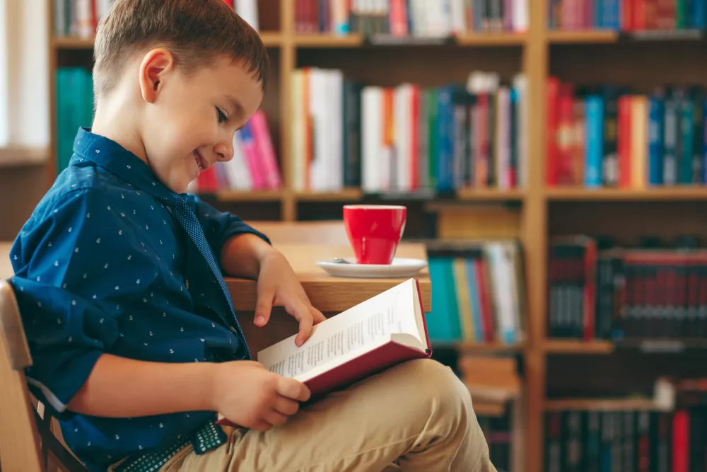 Boy reading a book sitting on a chair near a table with a cup of tea on the background of multi colored bookshelf in the library. Education, Knowledge, getting ready for school. / Foto: Diignat