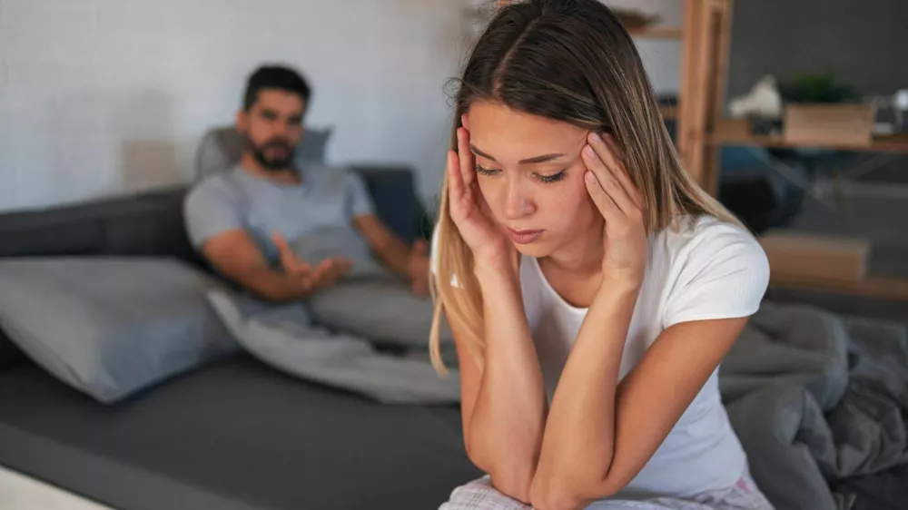 Beautiful depressed woman sitting on bed while her angry boyfriend yells at the background / Foto: Jelena Danilovic