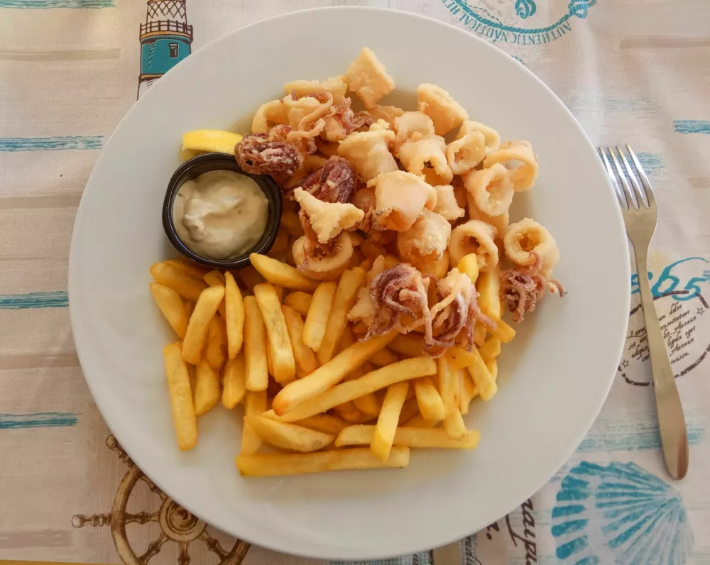 A white plate of fried potatoes, tiny octopus, squid rings and sauce on a light-coloured cloth napkin with a nautical motif. A metal fork lies next to it / Foto: Svetlana Barabanova