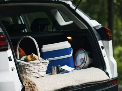 Open trunk of car showing picnic basket, cooler, badminton rackets, soccer ball, and travel gear packed for family road trip, outdoor forest background visible through trees