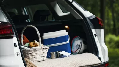 Open trunk of car showing picnic basket, cooler, badminton rackets, soccer ball, and travel gear packed for family road trip, outdoor forest background visible through trees