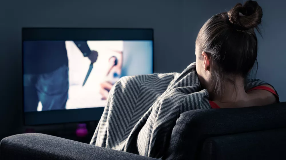 Watching horror and scary film on tv. Woman and thriller movie on television. Person in fear on home sofa couch under blanket at night in dark. Streaming true crime series or halloween show.