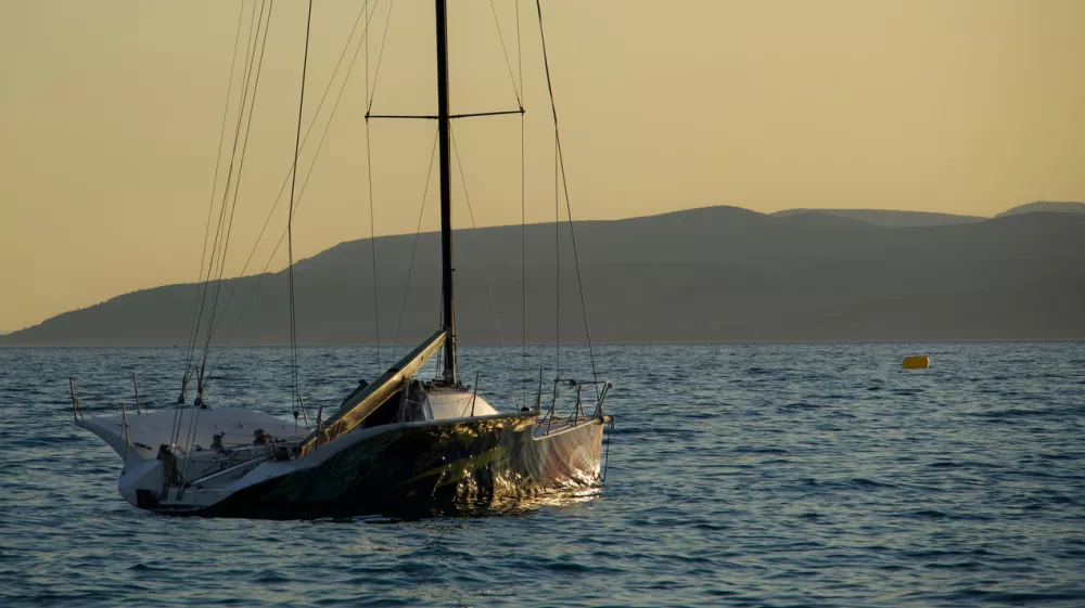 Small sailing yacht floating on gentle waves during golden sunset light. Peaceful seascape with distant mountains and warm evening atmosphere.