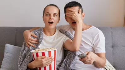Portrait of impressed woman watching horror film with man, shocked wife covered husband's eyes with palm, forbidden content on tv, couple sitting on sofa wrapped in blanket.