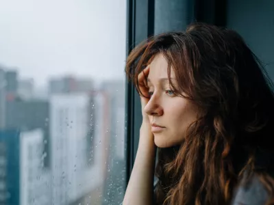 Young woman feeling sad and pensive, experiencing autumn depression while looking out a rainy city window, pondering loneliness and mental health challenges during a melancholic season