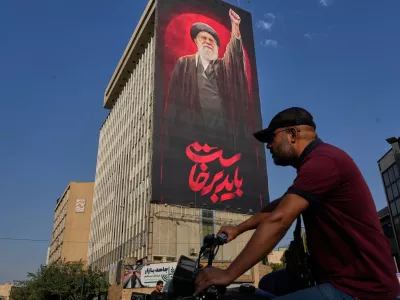 A man rides on his motorbike past a billboard with a message that reads in Farsi: " We must rise up" and an image of the late Iranian Supreme Leader Ayatollah Ali Khamenei, who was killed during the U.S. and Israel strikes on Feb. 28, in Tehran, Iran, Saturday, Aug. 15, 2026. (AP Photo/Vahid Salemi)