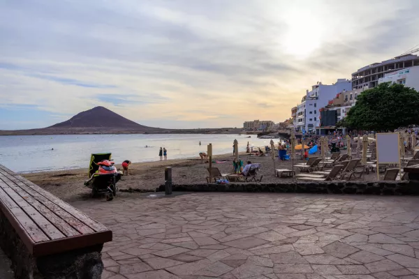 Beach with tourists, sunloungers, sunshades, Atlantic Ocean, hotels and volcanic cone Montana Roja (Red Mountain) at sundown in town El Medano on Canary Island Tenerife, Spain / Foto: Johannes86 Getty Images