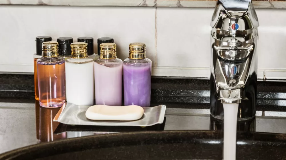 Water flows from the tap in the bathroom. Mini bottles of cosmetics on the table in the hotel room / Foto: Doroo