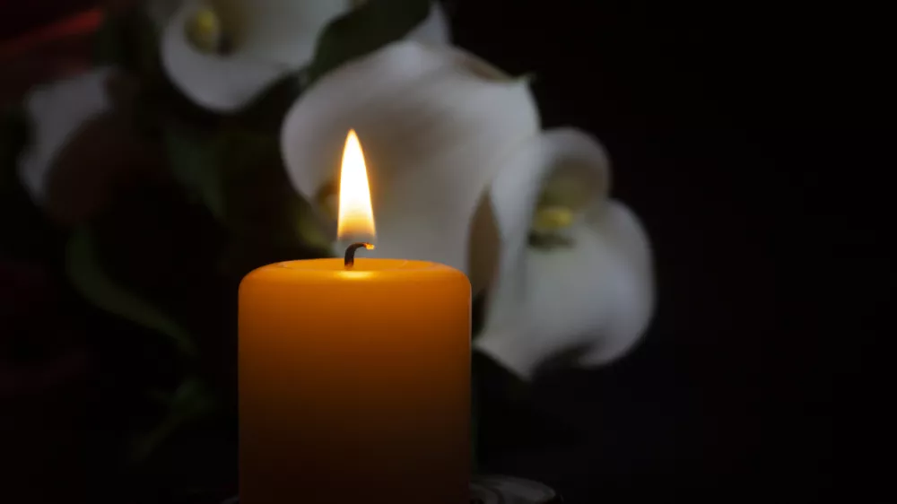 A close up of an orange candle and flame and lily flowers on a dark background.