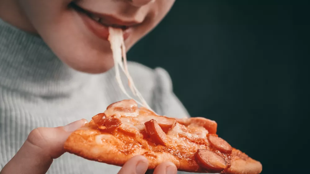 Happy caucasian girl holding a slice of pizza with stretching mozzarella cheese in her hand and biting her mouth on a black background in focus, close-up side view.