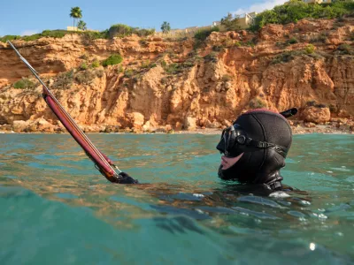 Side view of anonymous fisherman wearing diving mask and wetsuit swimming with speargun in blue sea surrounded by steep rocky shore in sunny day