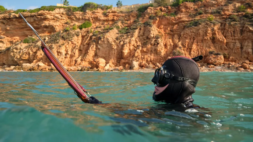 Side view of anonymous fisherman wearing diving mask and wetsuit swimming with speargun in blue sea surrounded by steep rocky shore in sunny day