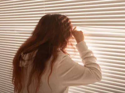 Rear view of beautiful young woman with long hair peeks through hole in the window blinds and looks out the window. Surveillance and curiosity