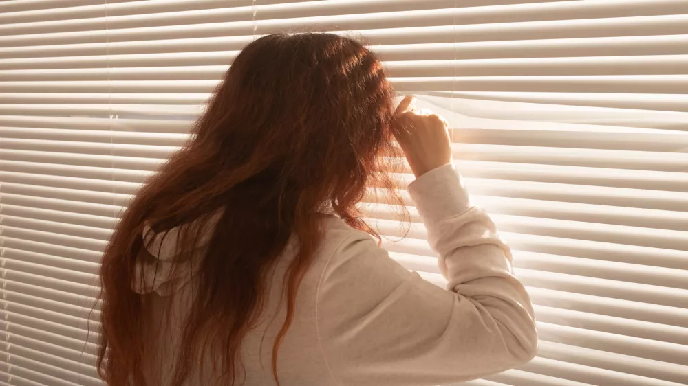 Rear view of beautiful young woman with long hair peeks through hole in the window blinds and looks out the window. Surveillance and curiosity