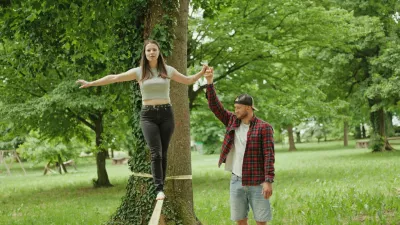 Young man helping woman balance on slackline by holding her hand in green park during outdoor recreation activity. Bright daylight, teamwork and focused movement create active Outdoors lifestyle and recreation atmosphere. / Foto: Logist Tehnik