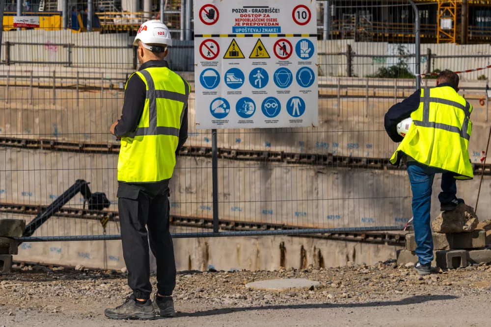 Ljubljana, Slovenia October 16, 2025 two construction workers wearing high‑visibility vests and safety helmets standing near a fenced‑off excavation area at an urban construction site, with multiple safety signs displayed, illustrating workplace safety, engineering, and infrastructure development. / Foto: Ijordan Getty Images