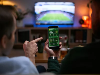 Young adult men watching football match and checking betting app on smartphone, expressing male friendship and shared leisure. Useful for sports betting, gaming, entertainment marketing