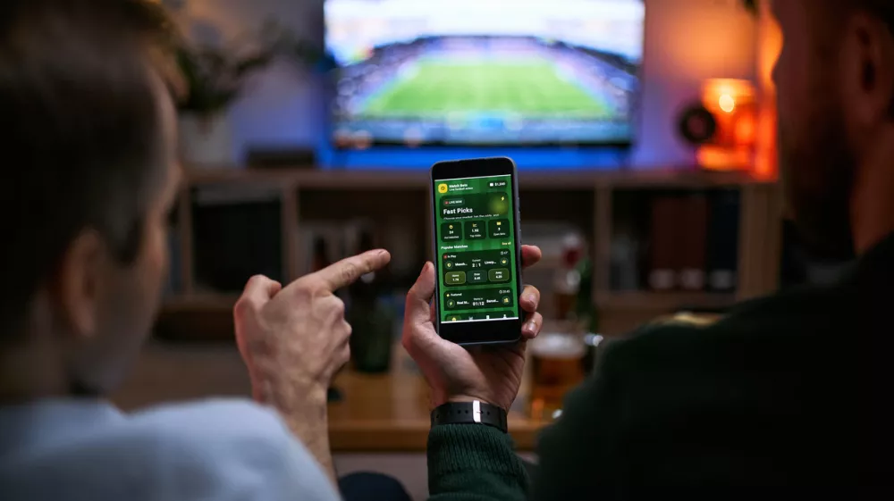 Young adult men watching football match and checking betting app on smartphone, expressing male friendship and shared leisure. Useful for sports betting, gaming, entertainment marketing