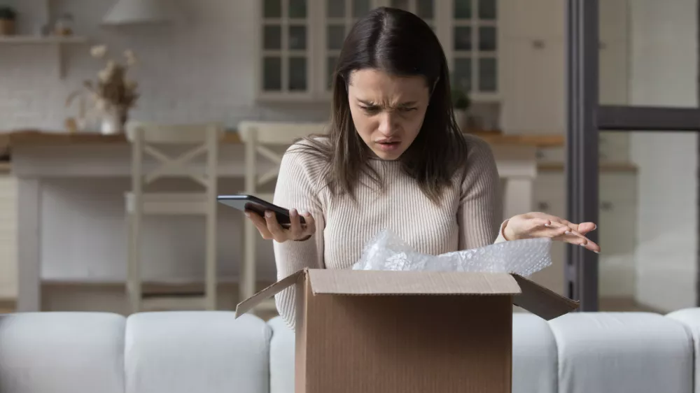 Puzzled dissatisfied customer getting damaged good from Internet store, opening wrong parcel, finding mistake. Confused worried young woman holding smartphone, checking carton box