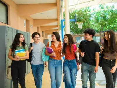 Group of cheerful latin teenage students walking together in school hallway and carrying backpacks and notebooks