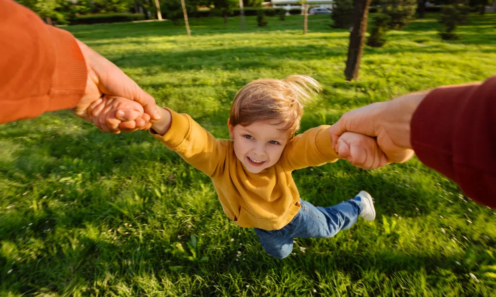 Smiling young boy holding hands with father while spinning playfully in a grassy park on a sunny day, full of excitement. Happy child portrait in movement during playtime. POV view
