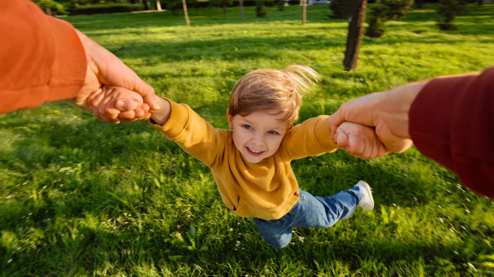 Smiling young boy holding hands with father while spinning playfully in a grassy park on a sunny day, full of excitement. Happy child portrait in movement during playtime. POV view