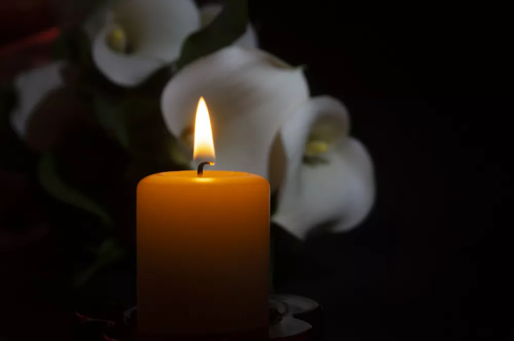 A close up of an orange candle and flame and lily flowers on a dark background.