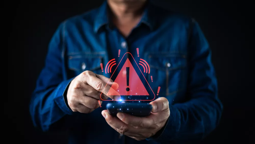 A person using a smartphone triggers a glowing red warning alert icon with an exclamation mark, symbolizing cybersecurity risks, data breaches, system errors, and mobile security threats.