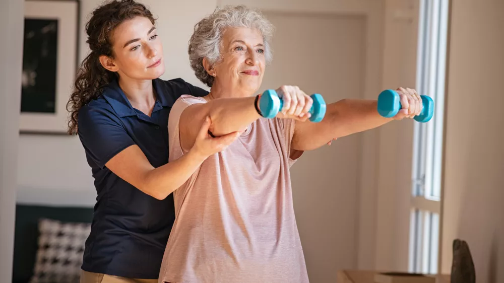 Old woman training with physiotherapist using dumbbells at home. Therapist assisting senior woman with exercises in nursing home. Elderly patient using dumbbells with outstreched arms in a physical therapy session in clinic.