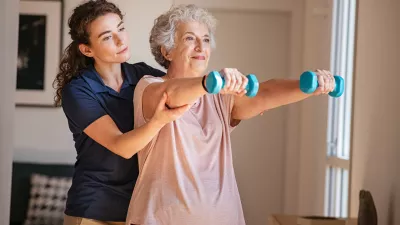 Old woman training with physiotherapist using dumbbells at home. Therapist assisting senior woman with exercises in nursing home. Elderly patient using dumbbells with outstreched arms in a physical therapy session in clinic.