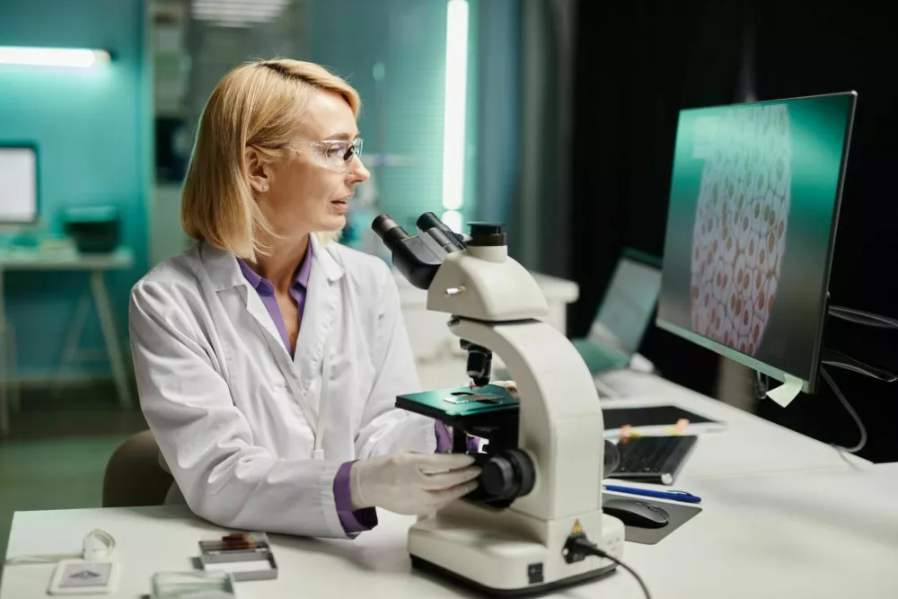 Caucasian middle aged female scientist examining biological samples under microscope while analyzing cell images on computer monitor in scientific laboratory, wearing protective eyewear and gloves / Foto: Seventyfour